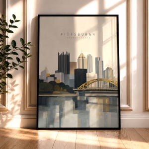 Op de afbeelding: Een ingelijste wandkunstprint met een gestileerde illustratie van de skyline van Pittsburgh, Pennsylvania. De afbeelding toont hoge gebouwen, een opvallende gele boogbrug en hun reflecties in het water, met de tekst PITTSBURGH PENNSYLVANIA in het midden bovenaan.