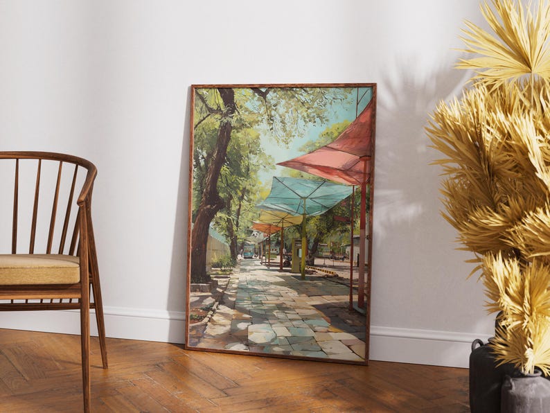 Puede incluir: Una pintura enmarcada representa una escena de calle soleada con toldos coloridos. La obra de arte presenta un camino de piedra bordeado de &aacute;rboles y edificios, con toldos rojos, azules y amarillos. La pintura se exhibe sobre un suelo de madera.