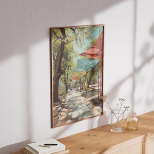 Puede incluir: Una pintura de acuarela enmarcada representa un camino empedrado soleado bordeado de &aacute;rboles y toldos coloridos. La obra de arte presenta una paleta de colores c&aacute;lidos con tonos de verde, rojo y azul. La pintura se exhibe en una pared blanca.