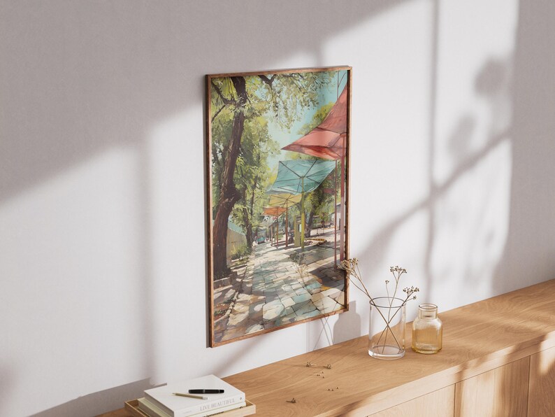 Puede incluir: Una pintura de acuarela enmarcada representa un camino empedrado soleado bordeado de &aacute;rboles y toldos coloridos. La obra de arte presenta una paleta de colores c&aacute;lidos con tonos de verde, rojo y azul. La pintura se exhibe en una pared blanca.