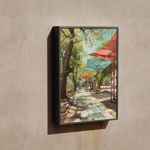 Puede incluir: Una pintura enmarcada representa una escena callejera soleada con toldos coloridos. La obra de arte muestra un camino adoquinado bordeado de &aacute;rboles y puestos de mercado, con toldos rojos, turquesas y amarillos. La pintura est&aacute; en un marco negro.