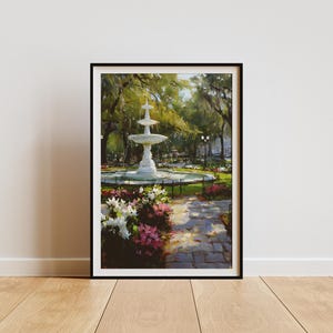 May include: Framed art print of a park scene featuring a tiered white fountain surrounded by flowers and trees. A stone path leads through the park. The artwork is in a black frame and is displayed on a white wall.