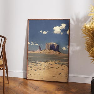 May include: Framed landscape art print featuring a desert scene with a large mesa under a blue sky with clouds. The artwork is in a wooden frame and leans against a white wall. A wooden chair with a yellow cushion is in the foreground.