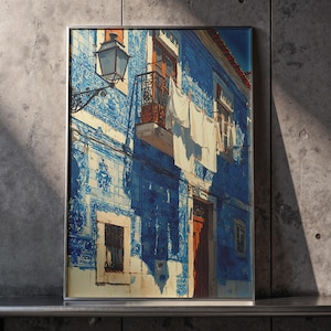 May include: Framed art print of a building facade with blue and white tilework. A balcony with wrought iron railings and hanging laundry is visible. A street lamp is mounted on the wall. The artwork is displayed against a concrete wall.