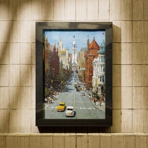 May include: A framed cityscape painting depicting a wide street lined with buildings in various architectural styles. The artwork features a blue sky and several cars, including yellow and white vehicles, on the road. The frame is black.