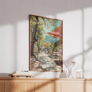 Puede incluir: Impresi&oacute;n art&iacute;stica enmarcada que representa una calle adoquinada soleada bordeada de &aacute;rboles y toldos coloridos. La obra de arte presenta una paleta de colores c&aacute;lidos con tonos de verde, rojo y amarillo. La impresi&oacute;n se exhibe en un mueble de madera.