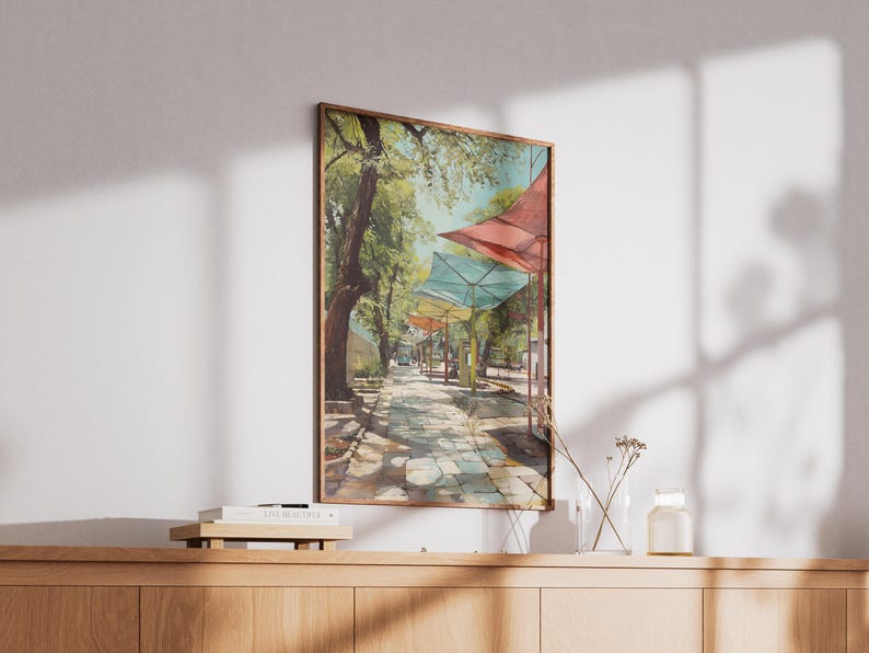 Puede incluir: Impresi&oacute;n art&iacute;stica enmarcada que representa una calle adoquinada soleada bordeada de &aacute;rboles y toldos coloridos. La obra de arte presenta una paleta de colores c&aacute;lidos con tonos de verde, rojo y amarillo. La impresi&oacute;n se exhibe en un mueble de madera.