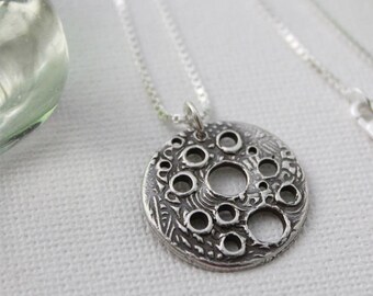 May include: A silver necklace with a round pendant featuring a textured design with multiple circles cut out. The chain is a delicate box chain.