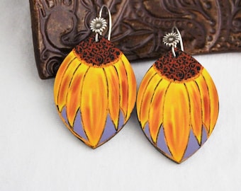 May include: A pair of handcrafted earrings featuring a sunflower design. The earrings have a yellow and orange petal design with a purple base and a dark brown center. The earrings are suspended from silver-colored hooks.