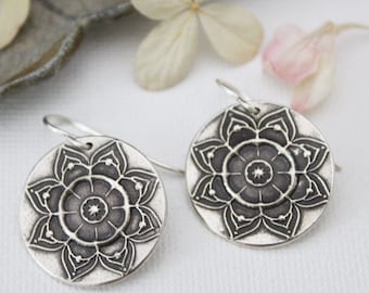 May include: A pair of silver dangle earrings with a mandala design. The earrings are round and have a detailed floral pattern.