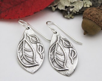 May include: A pair of silver earrings with a leaf design. Each earring features a teardrop-shaped pendant with three leaves embossed on it.