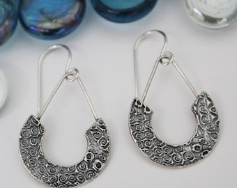 May include: A pair of silver earrings with a textured, swirl design. The earrings are shaped like a crescent moon and hang from a triangular wire.