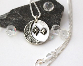 May include: A silver-toned pendant necklace featuring a crescent moon with etched details and two starbursts. The necklace hangs on a delicate chain. The pendant is displayed on a dark gray rock with clear crystal accents.
