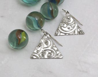 May include: A pair of silver triangle earrings with an ornate, swirling pattern. The earrings are displayed with several clear glass marbles with colorful swirls inside. The earrings have a hook closure.