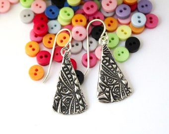 May include: A pair of silver dangle earrings with a triangular shape and intricate, detailed designs. The earrings are hanging against a background of colorful buttons.