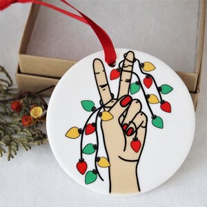 May include: White ceramic Christmas ornament featuring a hand making the peace sign, adorned with a string of colorful Christmas lights. The ornament has a red ribbon for hanging and is presented with a gift box.