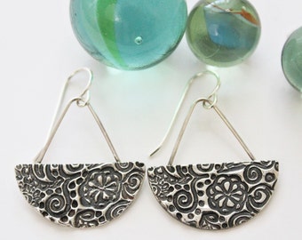 May include: A pair of silver earrings with a half-circle design featuring a detailed floral pattern. The earrings have a simple wire hook closure.