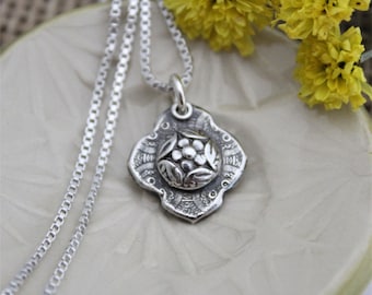 May include: A silver necklace with a delicate chain and a pendant featuring a floral design. The pendant is intricately detailed with a central flower and surrounding leaves.