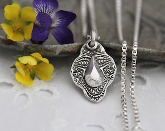 May include: A silver necklace with a pendant featuring a textured, ornate design and a small, teardrop-shaped charm. The necklace is on a white surface with yellow and purple flowers.