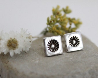 May include: A pair of silver earrings with a square shape and a flower design in the center.