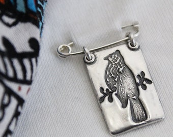 May include: A silver-toned brooch featuring a detailed bird design on a rectangular plaque. The bird is perched on a branch, and the brooch has a safety pin closure. The background is a white fabric.