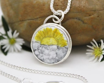 May include: A silver necklace with a round pendant featuring a hand-painted sunrise over clouds. The pendant is set in a silver bezel and has a silver chain.