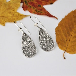 May include: A pair of silver teardrop earrings with a swirling, textured design. The earrings are hanging from a white surface.