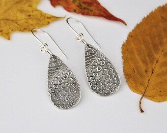 May include: A pair of silver teardrop earrings with a swirling, textured design. The earrings are hanging from a white surface.