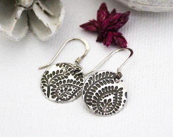 May include: A pair of silver-toned, round earrings with a detailed, etched leaf pattern. The earrings have a hook closure and are displayed on a white surface, with a dark red flower and a white object in the background.