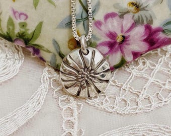 May include: A silver pendant necklace featuring a round, textured pendant with a floral design. The pendant hangs from a delicate silver chain. The background includes a floral pattern and lace.