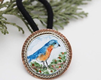 May include: A hair tie featuring a circular metal charm with a hand-painted blue and orange bird perched on green foliage. The charm has a hammered copper outer rim and a twisted silver-tone inner border surrounding the painted bird illustration.