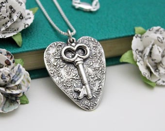 May include: A silver heart-shaped pendant necklace with an ornate key design. The pendant features intricate floral patterns and is suspended from a delicate chain. The necklace is displayed on a teal surface, with paper flowers in the background.