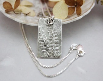 May include: A silver rectangular pendant necklace with a fern leaf design. The pendant is textured and hangs from a delicate silver chain. The necklace is displayed on a white surface with dried flowers.