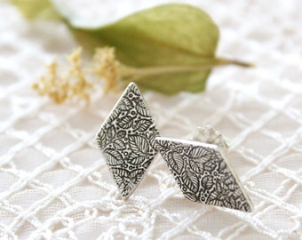 May include: A pair of silver earrings with a diamond shape and a leaf pattern. The earrings are on a white lace background.