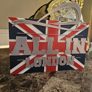 May include: A rectangular sign featuring a layered Union Jack flag design in red, white, and blue. The sign displays the silver-colored text "ALL IN" on the top line and "LONDON" on the bottom line, with a stylized silver arch graphic above.