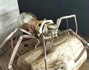 May include: A steampunk-style sculpture of a spider made from metal and a light bulb. The spider has a bulbous head, copper wire detailing, and articulated legs. It is mounted on a piece of weathered wood, creating a unique industrial art piece.