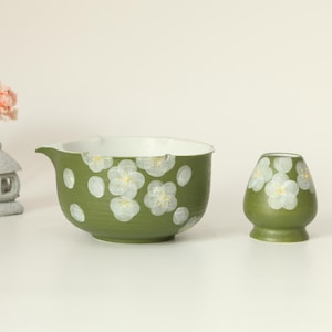 May include: A green ceramic tea bowl and matcha whisk holder, both decorated with white floral designs. A miniature pink cherry blossom tree and a stone lantern are also present, suggesting a Japanese theme.