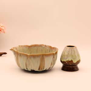 Puede incluir: Un cuenco de matcha de cerámica con un esmalte verde claro y marrón, un soporte para batidor a juego, una linterna de piedra decorativa y un cerezo en flor en miniatura. El cuenco tiene una forma única de pétalo.