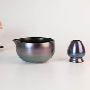 Puede incluir: Un cuenco de té negro e iridiscente con un pico, junto a un soporte para batidor a juego. Un pequeño árbol decorativo y una linterna de piedra están en el fondo. El cuenco y el soporte del batidor tienen un acabado nacarado.