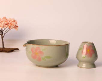 May include: A serene composition featuring a light green ceramic tea bowl and a matcha whisk holder, both adorned with pink floral designs. A miniature tree with pink blossoms and a stone lantern complete the scene, evoking a sense of tranquility.