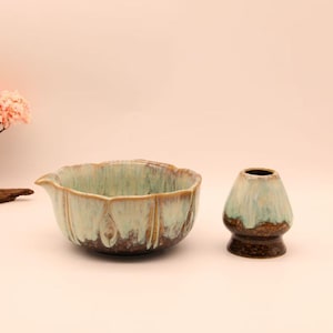 Op de afbeelding: Een keramische matcha kom, garde en een kleine decoratieve boom met roze bloesems. De kom heeft een lichtgroen en bruin glazuur. De garde is lichtgroen en bruin. De boom staat op een klein stukje hout.