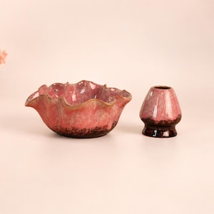 May include: A pink ceramic bowl with a ruffled, wavy rim and a matching pink ceramic whisk holder. Both items feature a glossy, mottled glaze with darker brown tones near the base, set against a plain background with a small decorative tree.