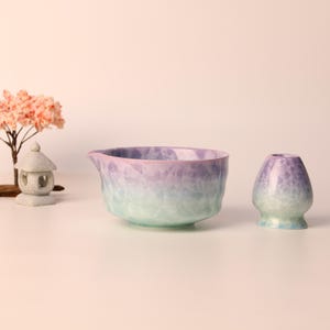 Puede incluir: Un cuenco de matcha y un soporte para batidor de cerámica en un degradado de morado y verde. Un pequeño árbol decorativo y una linterna de piedra están en el fondo. El cuenco tiene un pico.