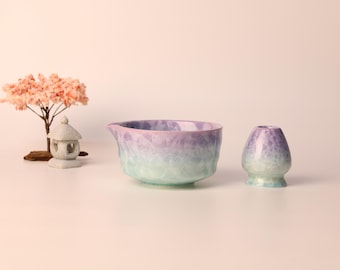 Può includere: Una ciotola per matcha e un porta frusta in ceramica in una sfumatura di viola e verde. Un piccolo albero decorativo e una lanterna di pietra sono sullo sfondo. La ciotola ha un beccuccio.