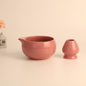 Può includere: Una ciotola per matcha in ceramica color rosa polvere con un piccolo beccuccio e un supporto per frusta in ceramica abbinato. Sullo sfondo, un piccolo albero di ciliegio decorativo e una lanterna in pietra in miniatura sono visibili.