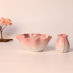 May include: A ceramic bowl and vase set with a pink and white gradient. The bowl has a ruffled edge, and the vase is shaped like a flower bud. A miniature tree with pink blossoms and a stone lantern are also present.
