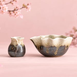 May include: A small ceramic sake carafe and a matching wide-mouthed pouring bowl. Both pieces feature a two-tone glaze with a dark, textured charcoal base that transitions into a smooth, creamy off-white top with a rippled, organic rim.