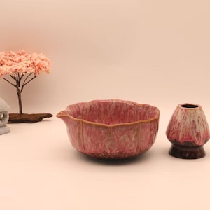 May include: A pink ceramic matcha bowl with a spout, a whisk holder, and a small decorative tree with pink blossoms. The bowl and whisk holder have a textured finish.