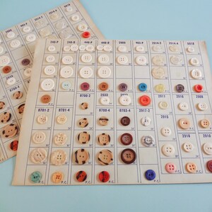 May include: Two vintage button cards with a variety of buttons in different sizes, colors, and materials. The buttons are arranged in rows and columns on the cards, with numbers and codes printed on the cards. The buttons include white, brown, red, and blue.