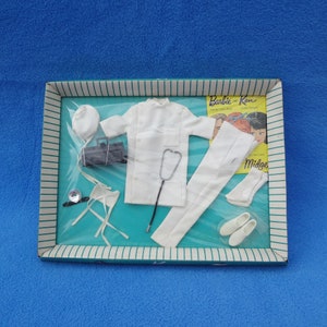 Puede incluir: Un conjunto de médico blanco para una muñeca Barbie, que incluye una bata de laboratorio, pantalones, sombrero, estetoscopio y una pequeña bolsa médica negra. El conjunto está empaquetado en una caja con una imagen de Barbie y Ken en la parte delantera.